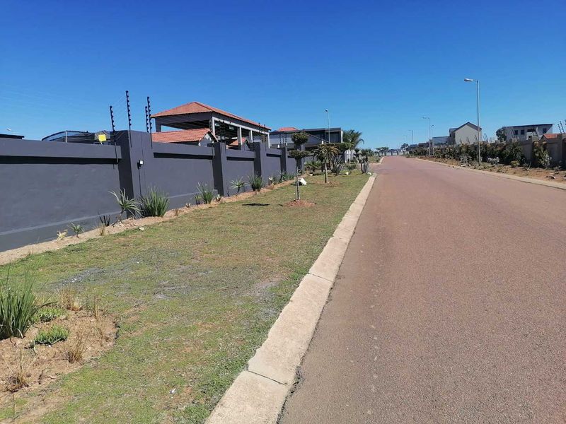 A clear street view of a residential area featuring paved roads, concrete curbing, and secure perimeter walls with electric fencing.