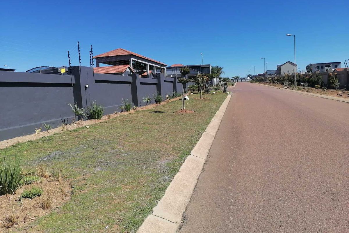 A clear street view of a residential area featuring paved roads, concrete curbing, and secure perimeter walls with electric fencing.