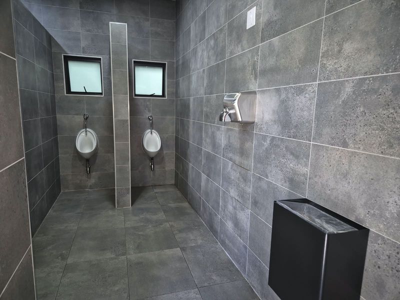 Modern commercial restroom featuring grey tiled walls and floors, equipped with wall-mounted urinals and a stainless steel hand dryer.