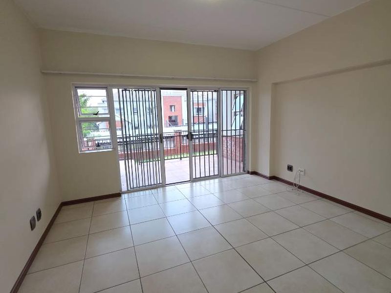 A bright, empty living room featuring neutral tiled flooring, a ceiling light fixture, and a sliding glass door with a security gate leading outside.