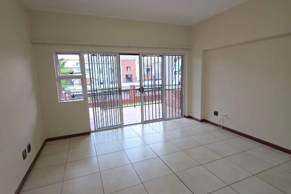 A bright, empty living room featuring neutral tiled flooring, a ceiling light fixture, and a sliding glass door with a security gate leading outside.