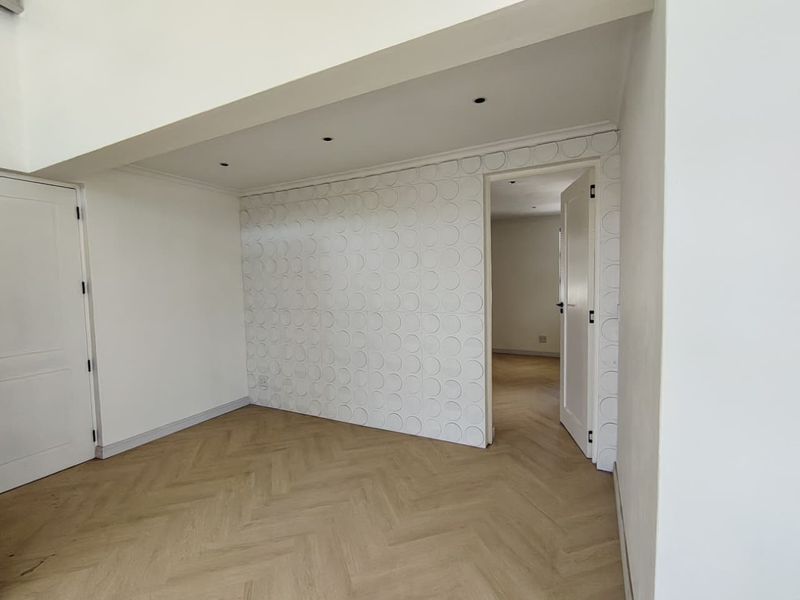 Bright, empty living area featuring modern herringbone flooring, recessed downlights, and a unique textured feature wall, leading into an adjacent room.