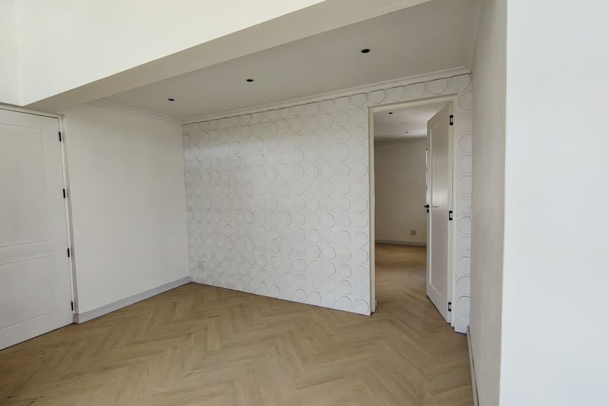 Bright, empty living area featuring modern herringbone flooring, recessed downlights, and a unique textured feature wall, leading into an adjacent room.