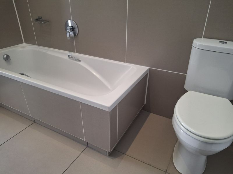 A clean, modern bathroom featuring a built-in bathtub with chrome fixtures and a standard ceramic toilet, finished with neutral wall and floor tiling.
