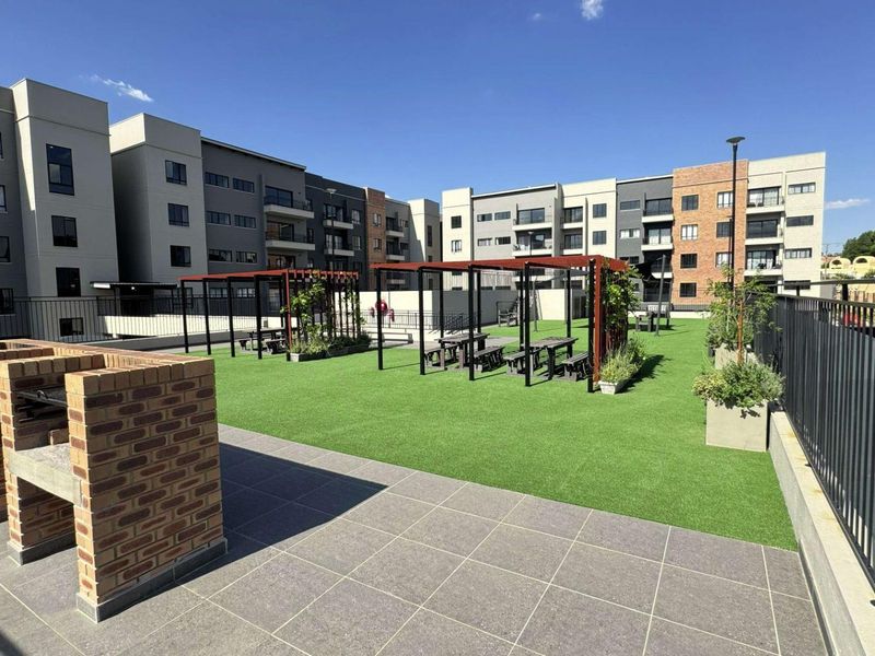 Communal outdoor entertainment area featuring a built-in brick braai, pergola-covered seating spaces, and well-maintained artificial lawn within an apartment complex.