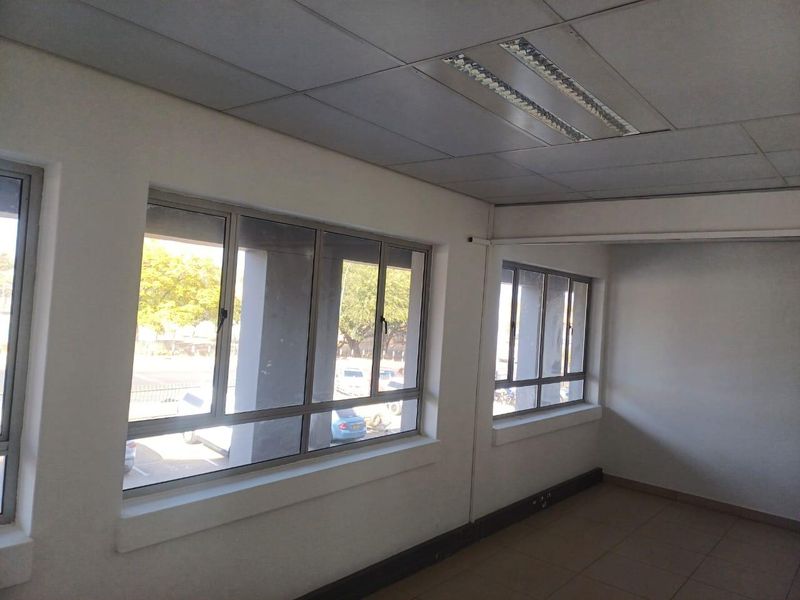 A bright, empty office space featuring tiled flooring, suspended ceilings with recessed lighting, and large aluminium-framed windows providing natural light.