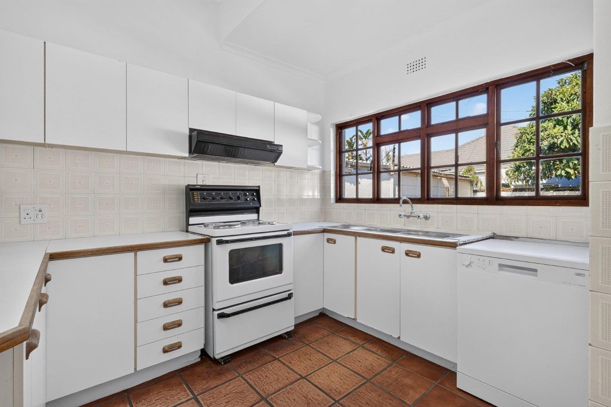 A functional kitchen featuring white cabinetry, a freestanding stove with extractor fan, double sink, and classic terracotta floor tiles with natural light.