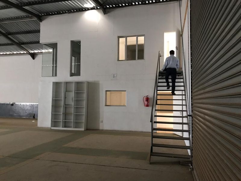 Industrial warehouse space featuring a mezzanine level with office access, metal staircase, roller shutter door, and exposed steel roof structure.