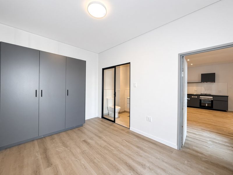 A modern, empty bedroom featuring sleek grey built-in cupboards, light wood-look laminate flooring, and an en-suite bathroom access point.