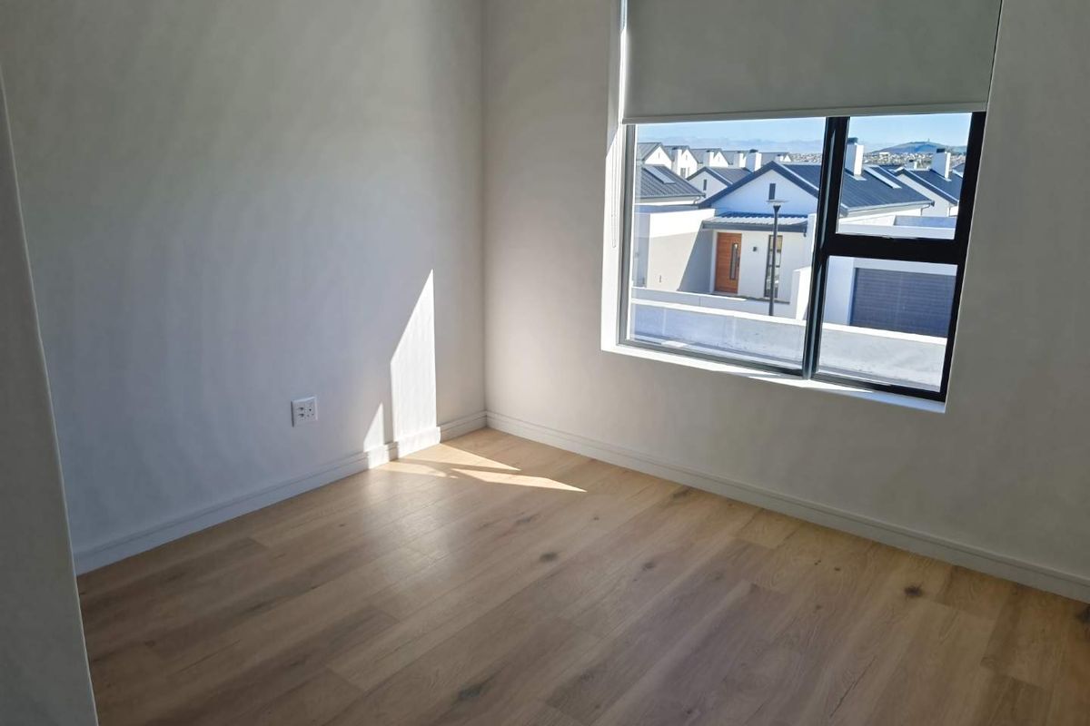 A bright, empty bedroom featuring modern light wood-look flooring, neutral walls, and a large window with a roller blind offering a neighbourhood view.