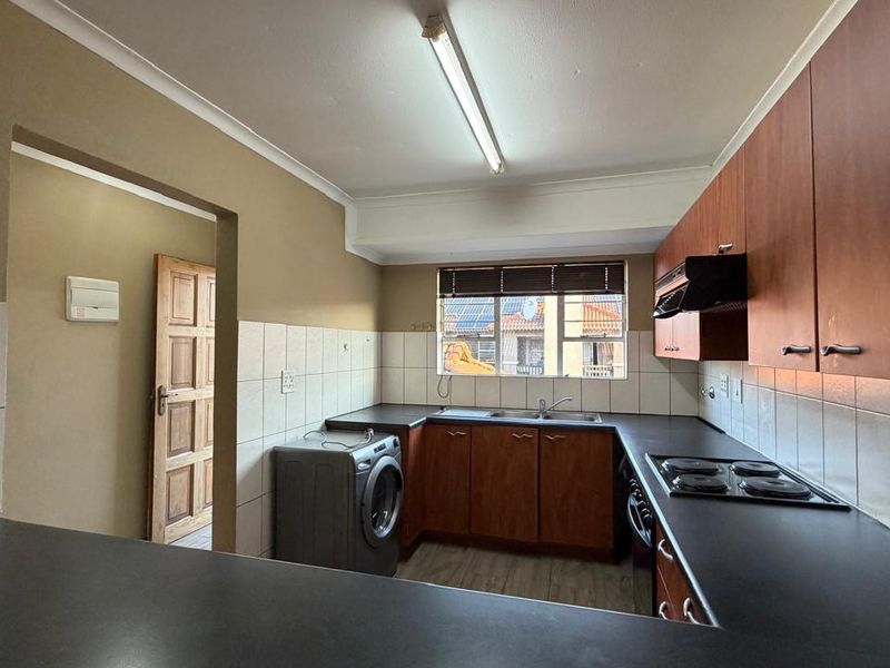 A functional kitchen featuring wood-finish cabinetry, dark laminate countertops, a built-in electric hob, and a tiled backsplash with integrated laundry appliance space.