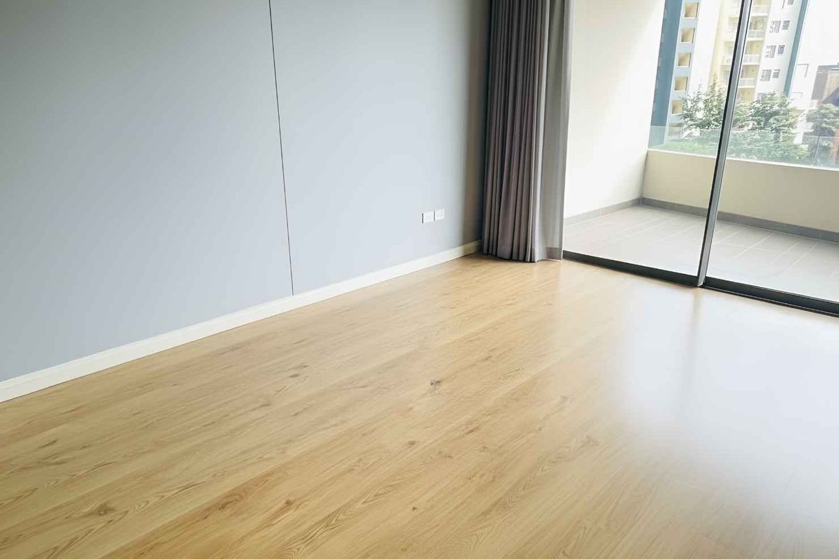 A bright, empty bedroom featuring modern laminate flooring, neutral wall finishes, and a large sliding glass door providing access to a private balcony.