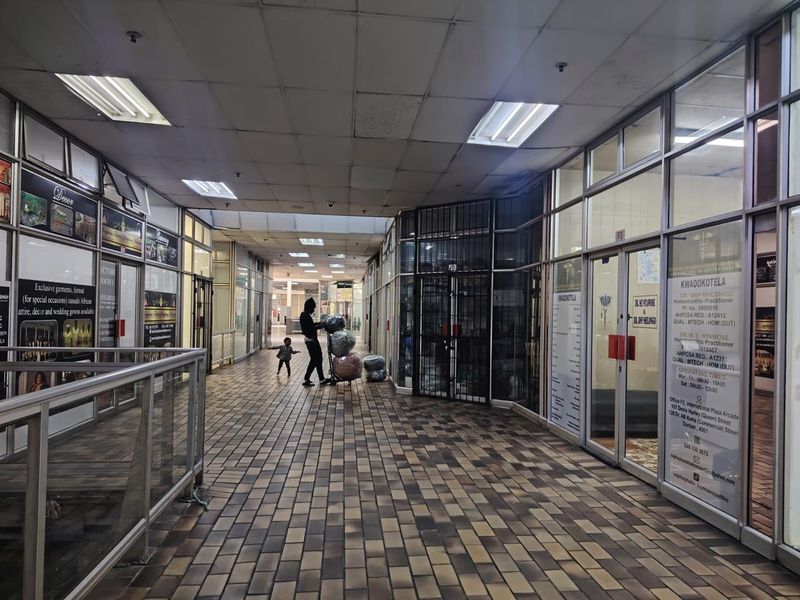 A commercial hallway in a retail plaza featuring tiled flooring, suspended ceiling panels, and multiple glass-fronted shop units with metal security frames.