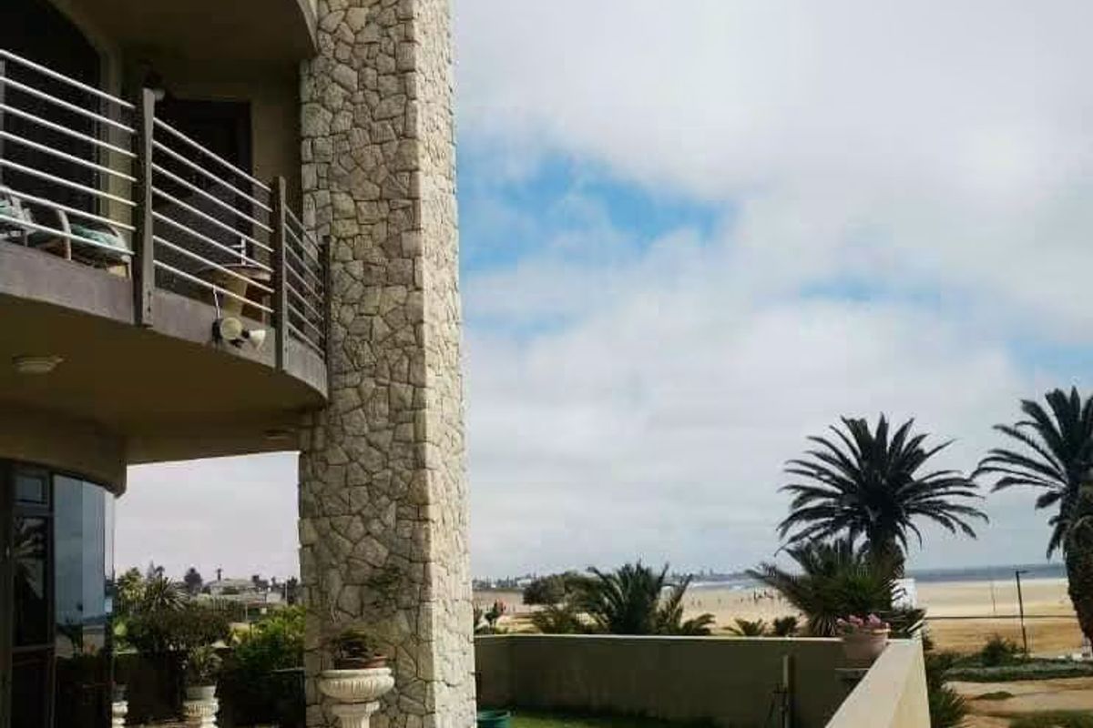 Rear exterior view featuring a stone-clad chimney, tiled patio, balcony with metal railings, and a landscaped lawn overlooking the beach.