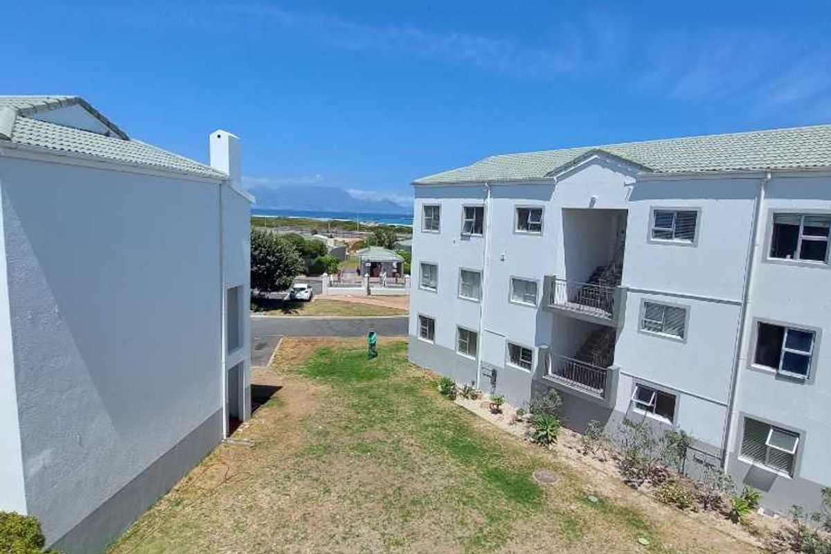 Exterior view of apartment complex buildings featuring tiled roofs, balconies, and a shared lawn area with distant coastal views.