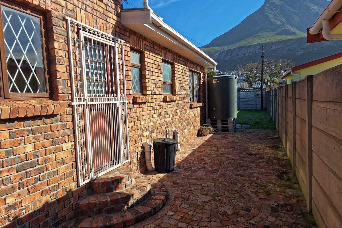 A side exterior view of the brick house featuring a security gate, paved walkway, and a water storage tank with mountain views.