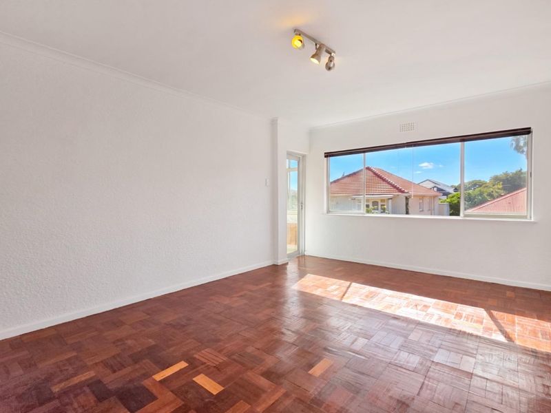 A bright, empty living room featuring classic parquet flooring, track lighting, and large windows that provide natural light and a pleasant neighborhood view.