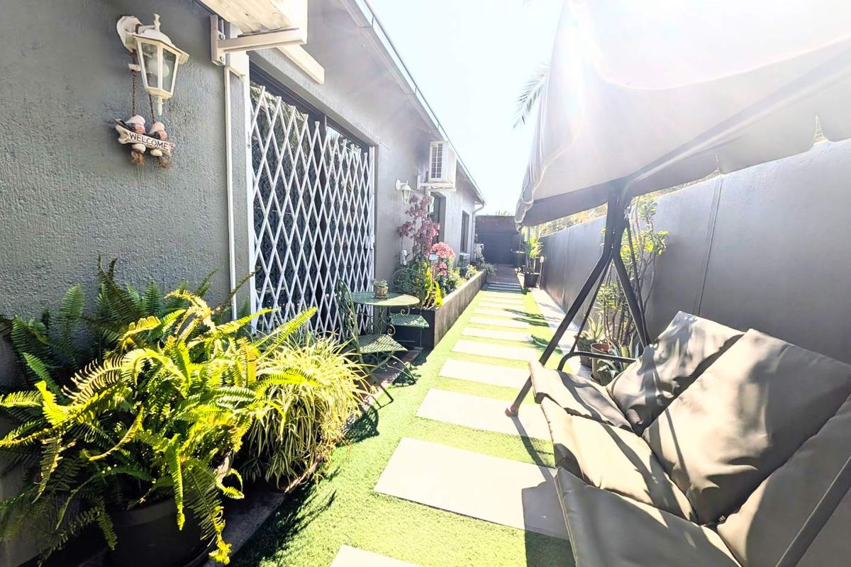 A narrow side garden area featuring a paved walkway, artificial grass, security gate, and wall-mounted lighting along the exterior wall.