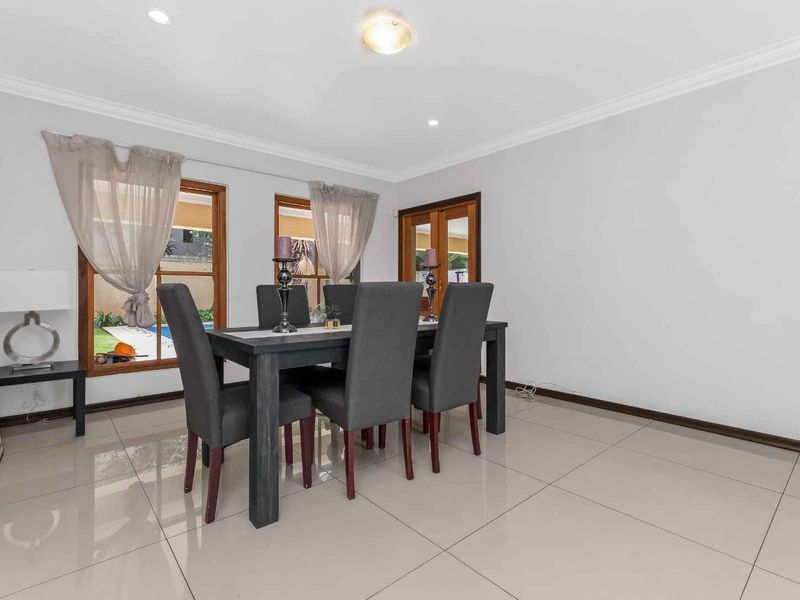 A bright dining room featuring large tiled floors, recessed lighting, and wooden-framed windows looking out toward the garden area.