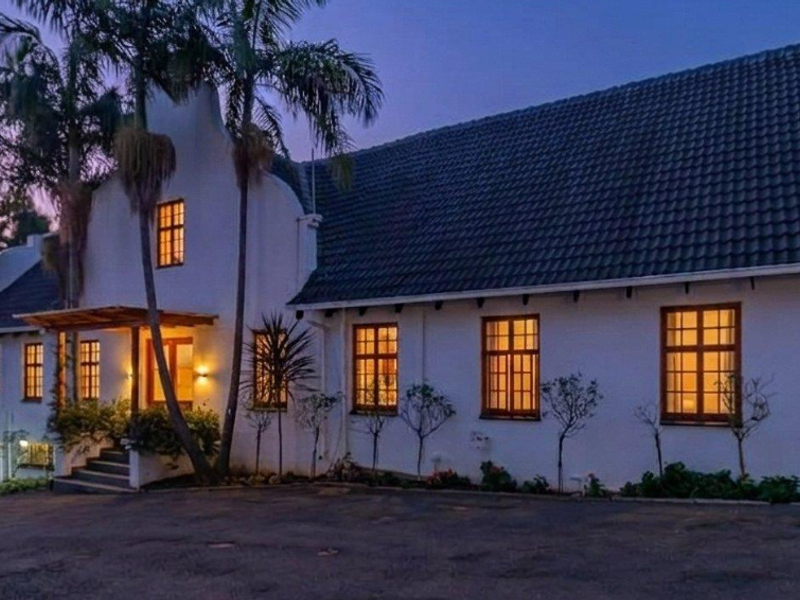 A charming Cape Dutch style home exterior at dusk, featuring a tiled roof, timber-framed windows, and a welcoming covered entrance porch.