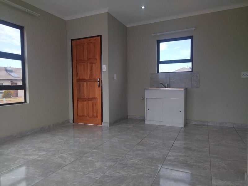 A clean, empty kitchen area featuring tiled flooring, a single basin sink with tiled splashback, and natural light from aluminium-framed windows.