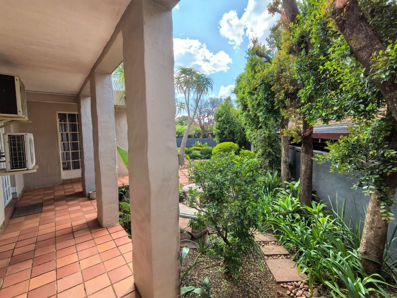 A covered patio area featuring terracotta tiling and concrete pillars, overlooking a lush, established garden with mature trees and a paved walkway.