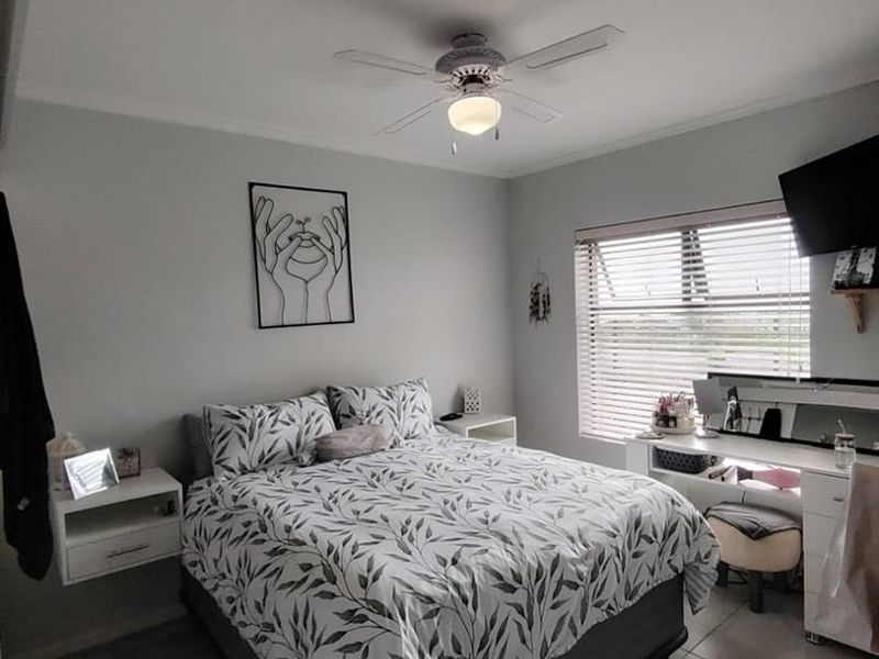 A well-maintained bedroom featuring neutral wall tones, tiled flooring, a central ceiling fan, and large windows fitted with venetian blinds.