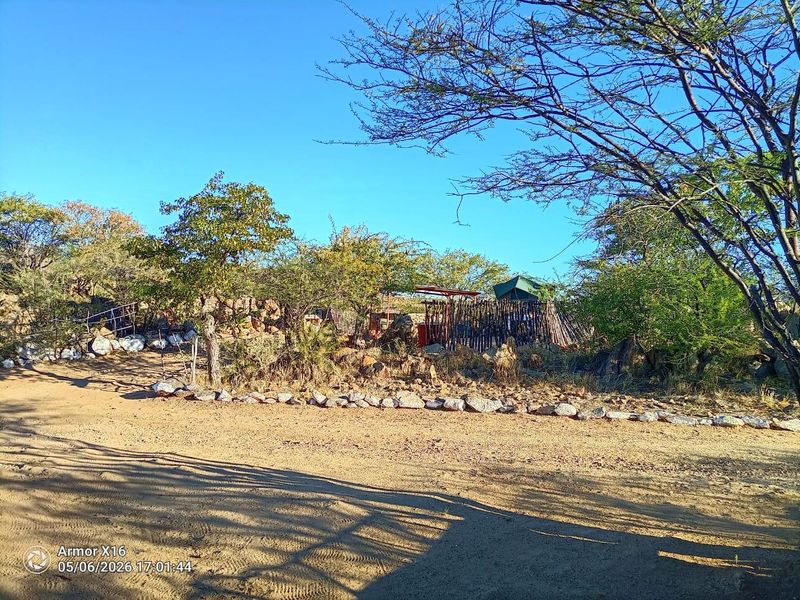 A rustic outdoor area featuring natural rock landscaping, wooden pole fencing, and a small thatched structure set within a natural bushveld environment.