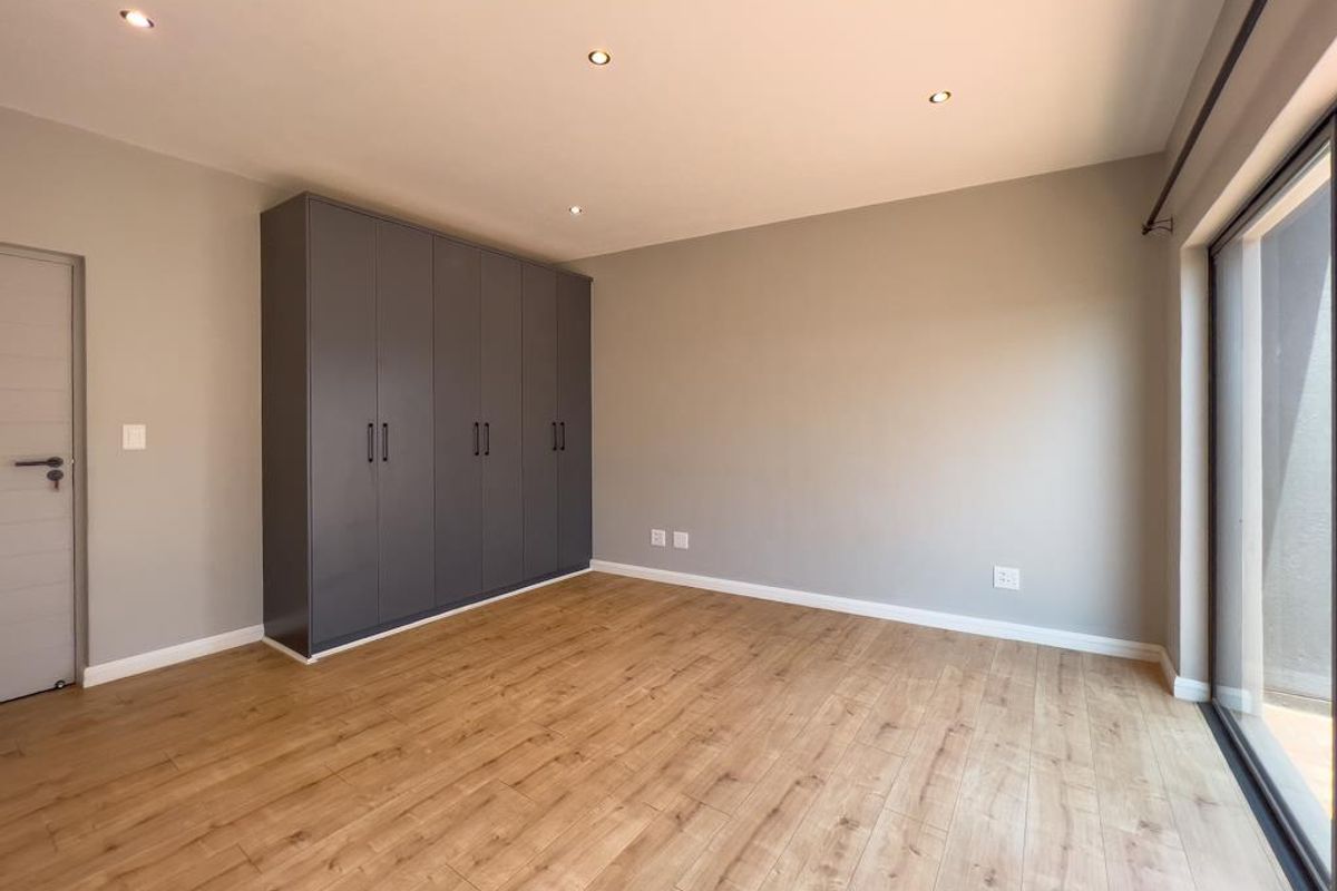 A bright, modern bedroom featuring light wood-look flooring, neutral walls, recessed ceiling lighting, and a large, dark grey built-in wardrobe.