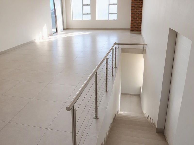 A bright, modern open-plan landing area featuring neutral tiled flooring, a sleek stainless steel balustrade, and an exposed brick accent column.