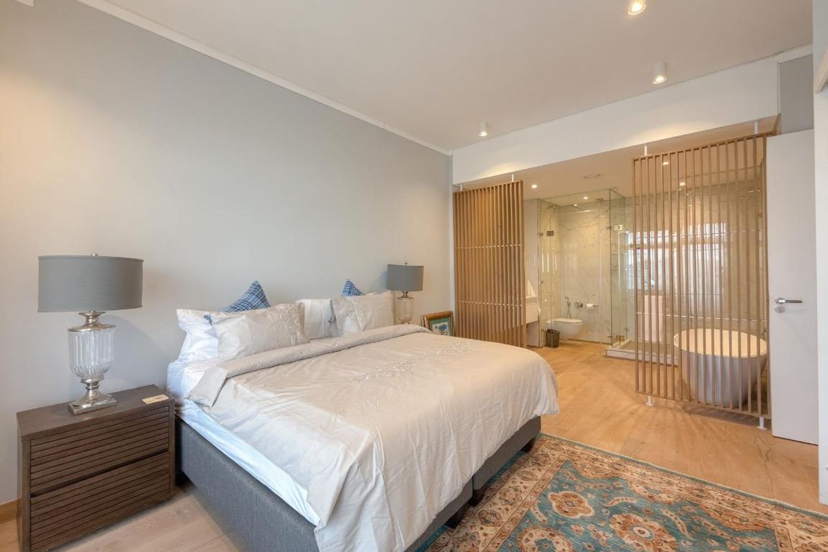 A modern bedroom featuring an open-plan ensuite with a freestanding bathtub, glass-enclosed shower, and a decorative wooden slat partition.