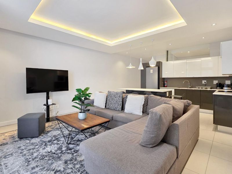 Modern open-plan living area featuring a sleek kitchen with island, recessed ceiling lighting, and neutral tiled flooring throughout the combined space.
