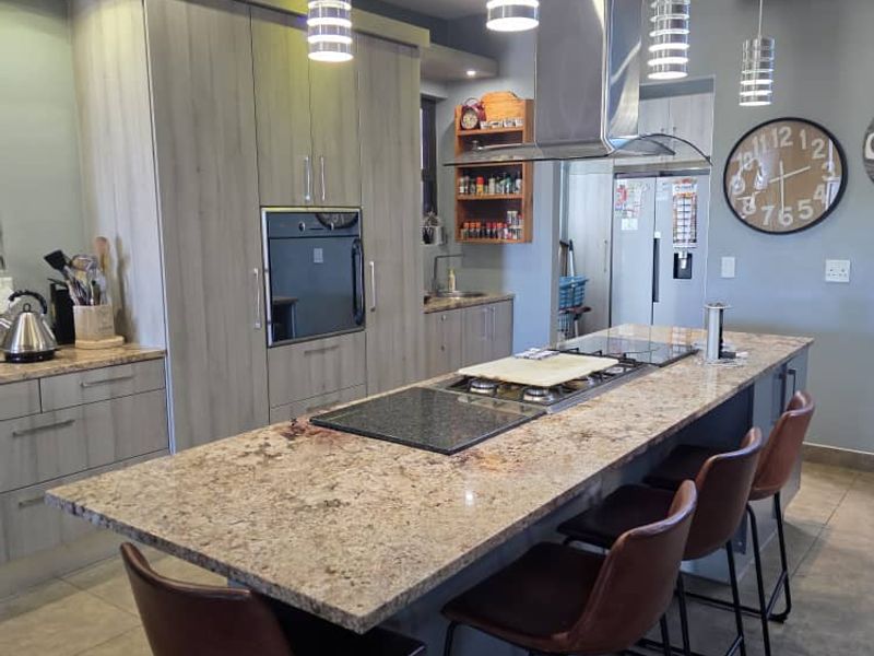 Modern kitchen featuring a large granite-topped island with integrated hob, stainless steel extractor fan, built-in oven, and contemporary cabinetry with recessed and pendant lighting.