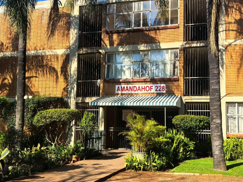 Front elevation of the Amandahof apartment complex, featuring a brick facade, security bars on windows, a covered entrance, and established garden landscaping.