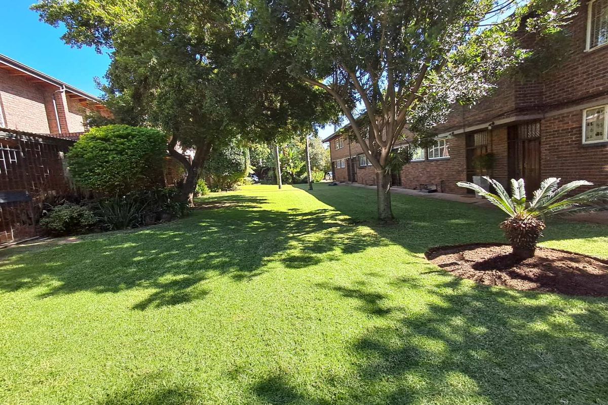 A well-maintained communal garden area featuring mature trees, manicured lawn, and brick townhouse facades.
