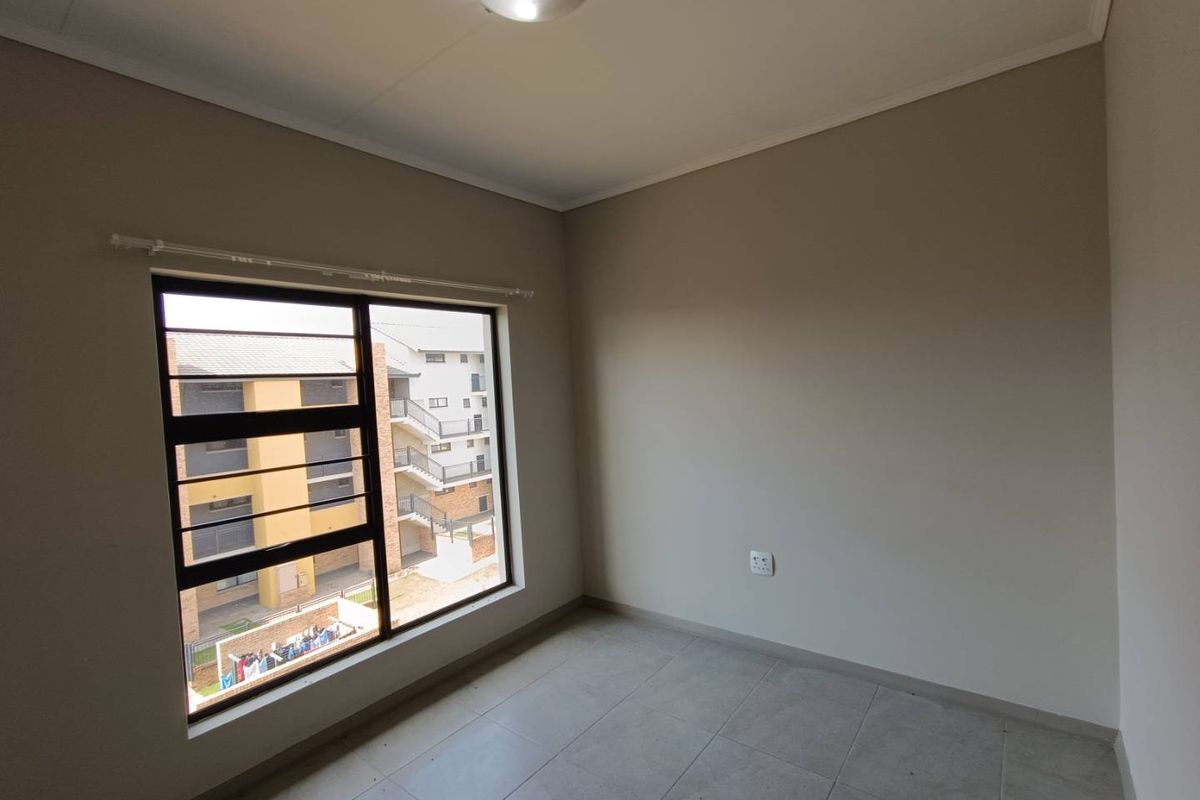 A bright, empty bedroom featuring neutral wall tones, tiled flooring, and a large window with security bars overlooking the apartment complex.