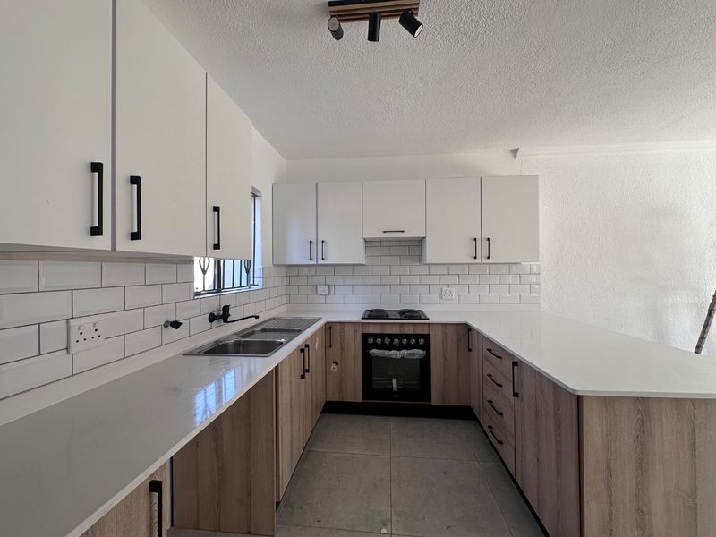 A modern, clean kitchen featuring white upper cabinets, wood-grain lower cabinetry, quartz countertops, subway tile backsplash, and integrated built-in oven and hob.