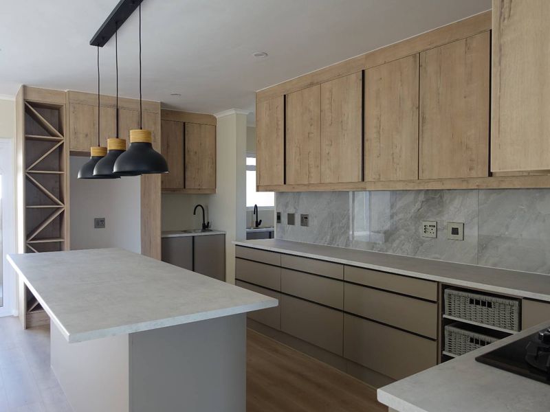 Modern kitchen featuring light wood-tone cabinetry, stone countertops, a central island, marble-look splashback, and integrated storage solutions in a clean, contemporary layout.