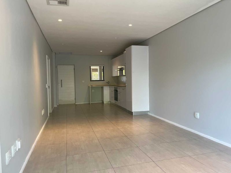 An open-plan living and kitchen area featuring modern white cabinetry, integrated appliances, neutral tiled flooring, and recessed lighting in a clean, vacant space.