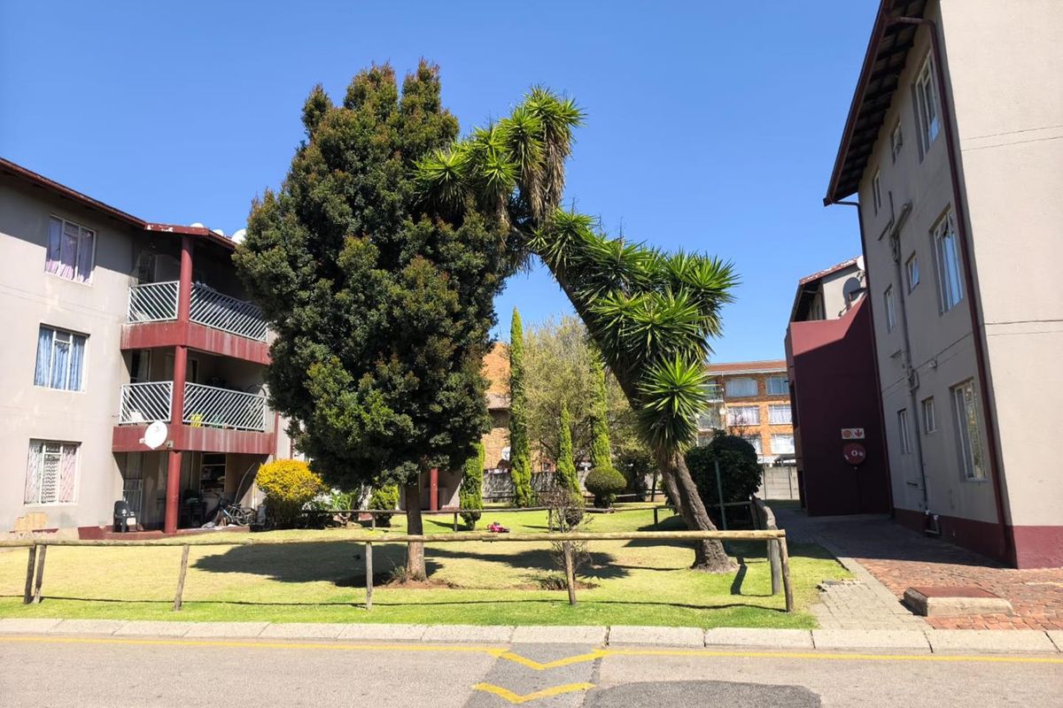A view of the apartment complex exterior featuring a shared garden area, paved walkways, and balconies overlooking the communal grounds.