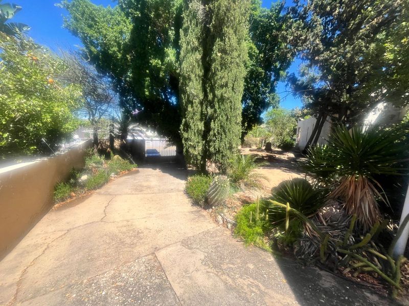 A front exterior view featuring a concrete driveway, mature trees, and established garden landscaping leading to a gated entrance.