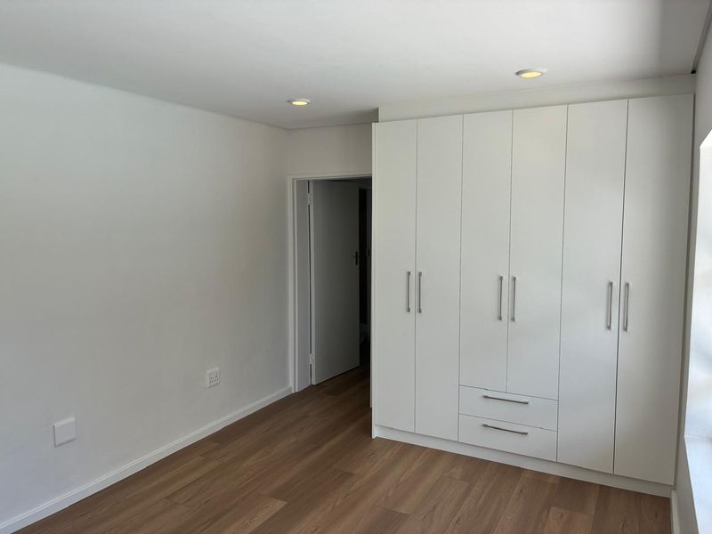 A bright, modern bedroom featuring clean white built-in cupboards, recessed ceiling lighting, and warm-toned laminate flooring.