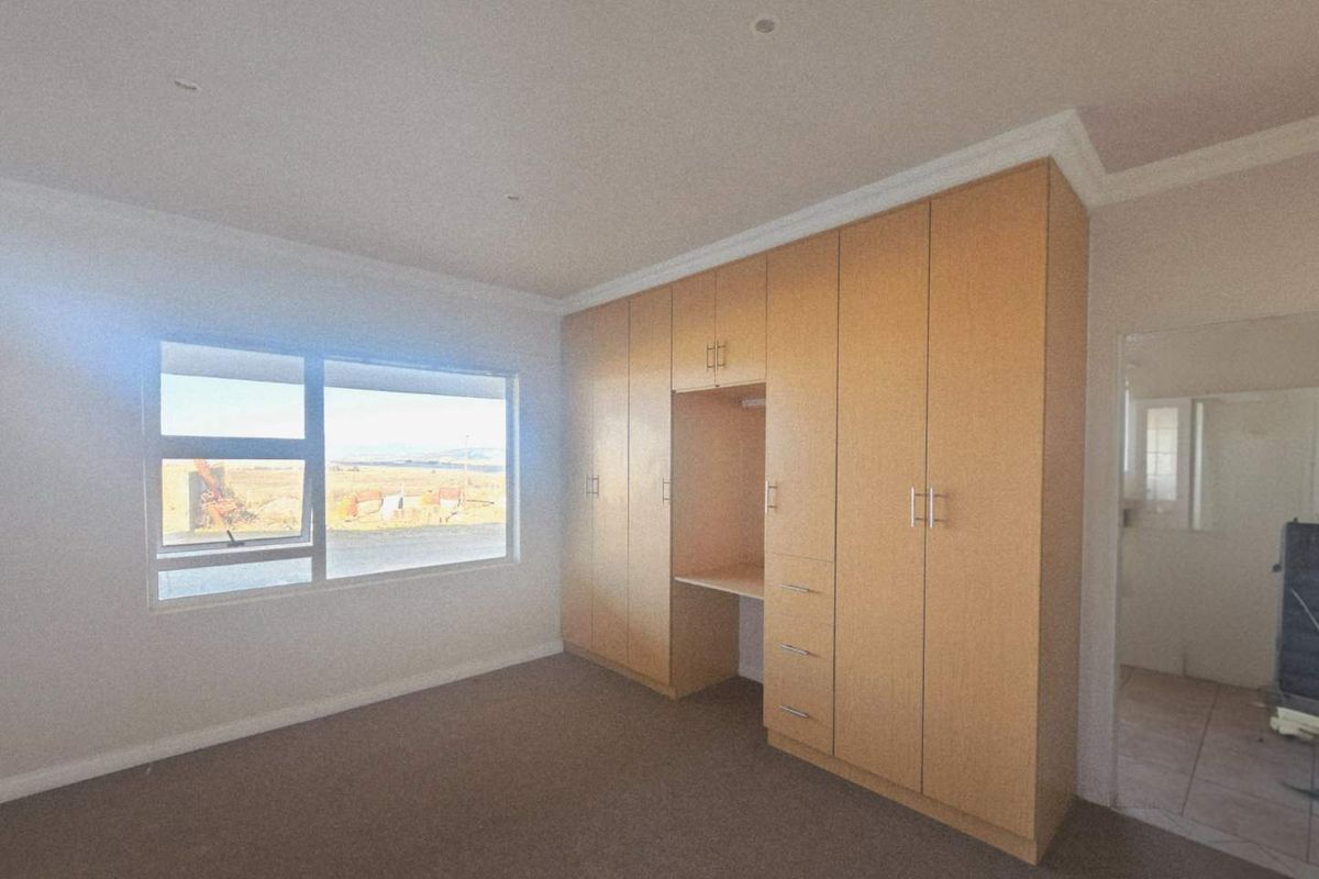 A spacious bedroom featuring built-in wooden cabinetry with an integrated desk area, neutral carpeting, and large windows offering natural light.