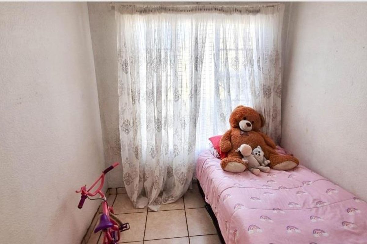A compact bedroom featuring tiled flooring and a window with sheer curtains, providing natural light to the space.