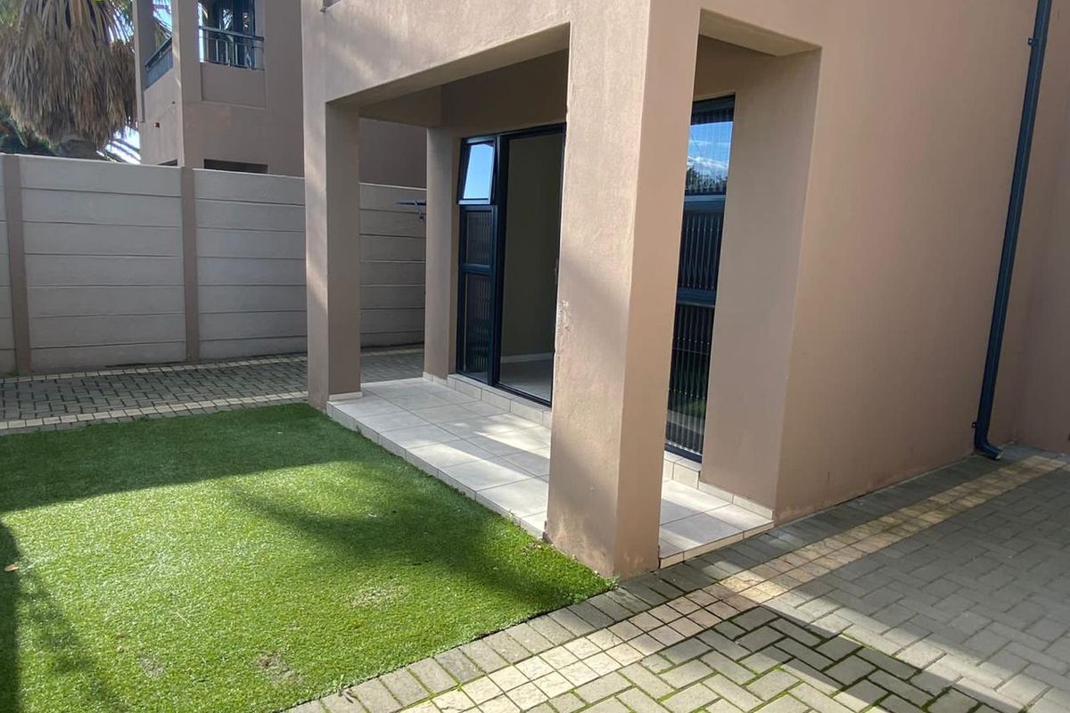 A rear exterior view of the apartment featuring a covered tiled patio, artificial lawn, and paved outdoor area enclosed by a boundary wall.