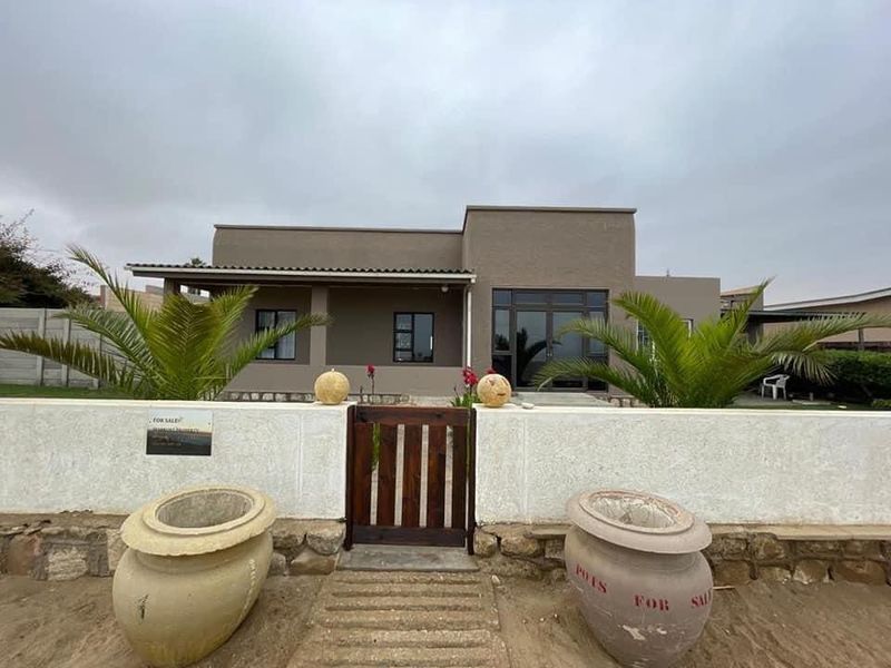 Front elevation of a modern single-story house featuring a covered patio, boundary wall with a wooden gate, and aluminium window frames.