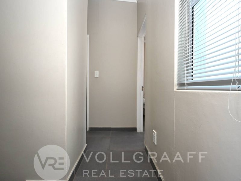 A clean, neutral-toned hallway featuring tiled flooring and a window fitted with venetian blinds, providing a bright and functional transition space.