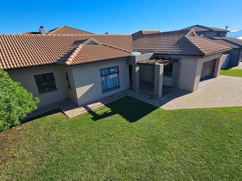 A well-maintained single-story house featuring a tiled roof, paved driveway, integrated garage, and a front pergola overlooking a manicured lawn.