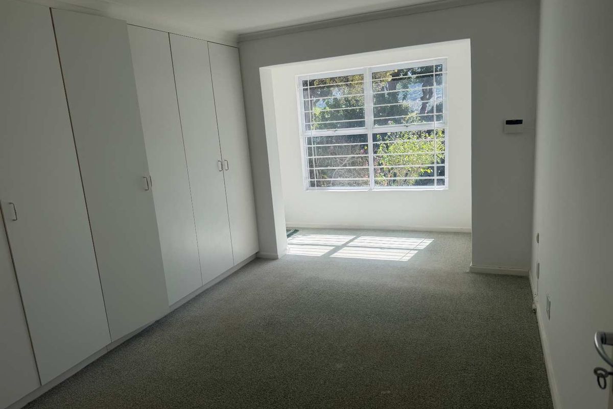 A bright, empty bedroom featuring wall-to-wall built-in cupboards, neutral carpeting, and a large window with security bars.