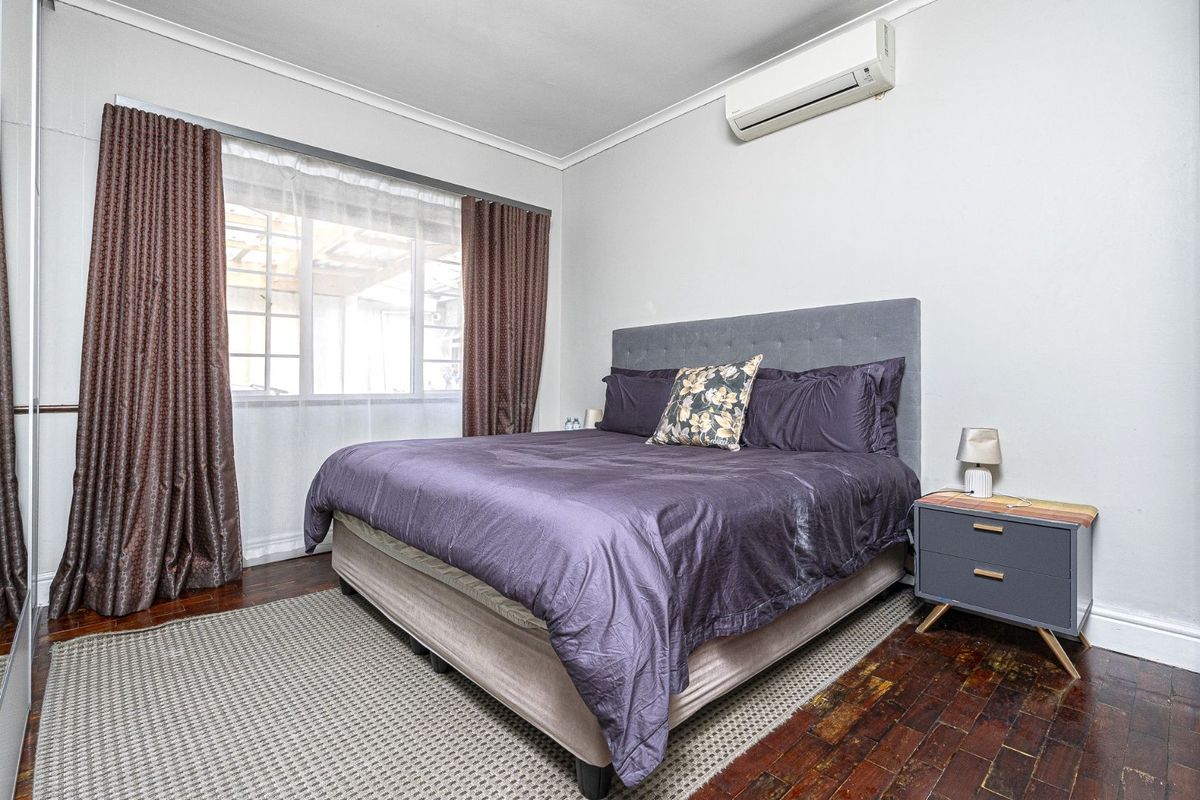 A well-lit bedroom featuring parquet flooring, a wall-mounted split-system air conditioner, and large windows with curtains.